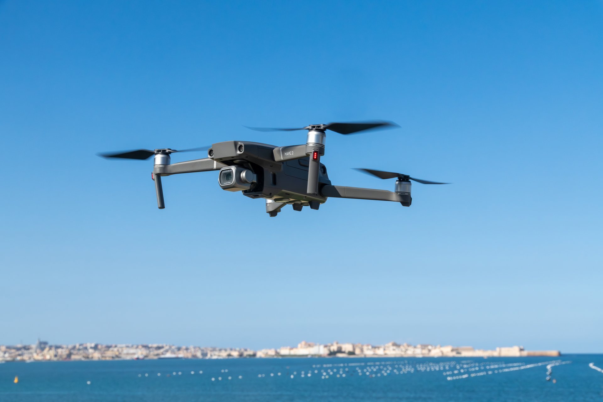 Drone hovering above coastal scene at sunset with spinning propellers, framed by calm sea and distant skyline under clear sky