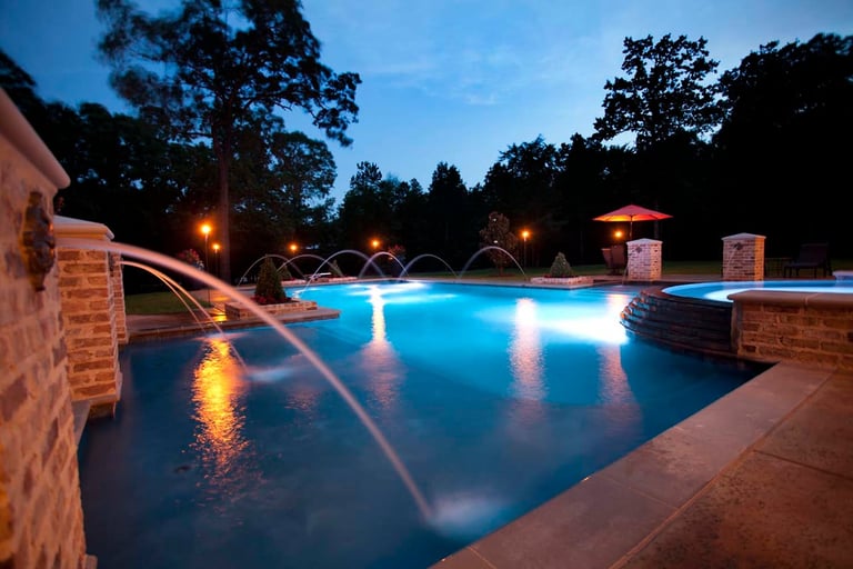 Illuminated swimming pool at dusk with glowing blue water, fountains, and landscape lighting surrounded by trees