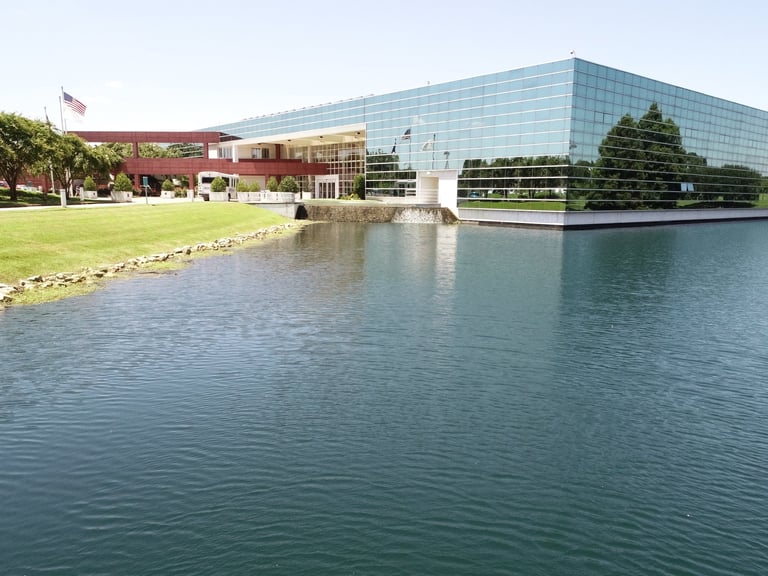Modern glass building with green roof beside calm water, landscaped grounds with trees and American flag