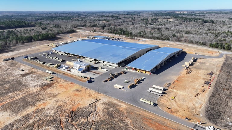 Aerial view of a large warehouse with bright blue roof panels and parking lot surrounded by bare trees and forest