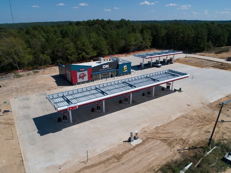 Aerial view of a commercial building under construction with metal frame structure and red brick sections, surrounded by trees and cleared land.