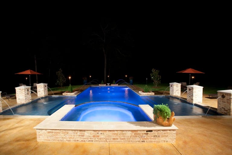 Luxurious backyard resort-style pool complex at night with blue illuminated main pool, spa jets, surrounding stone decking, lounge areas with umbrellas, and landscape lighting.