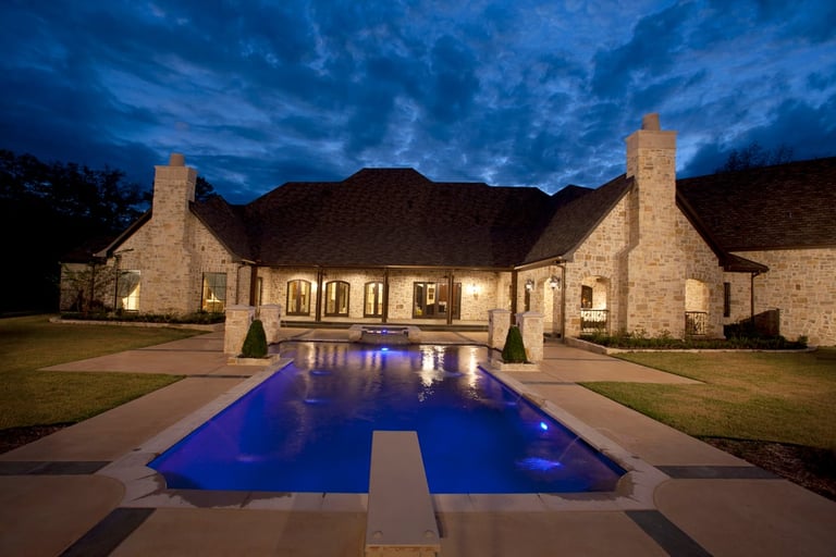 Illuminated stone mansion with blue lit swimming pool at dusk under dramatic evening sky