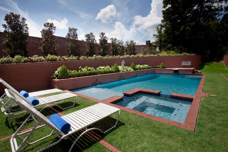 Backyard with rectangular swimming pool, hot tub, lounge chairs on grass, red brick walls with greenery, and clear blue sky