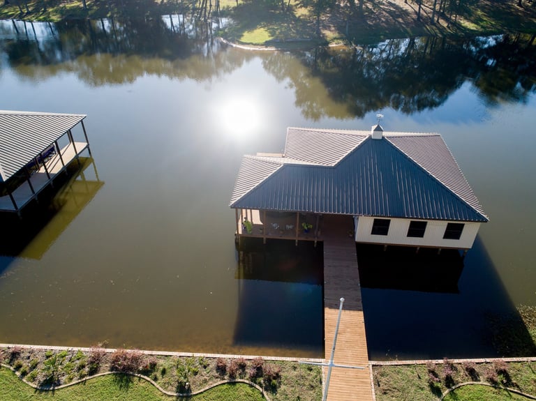 Aerial view of a boathouse with twin peaked roofs on a calm river, connected by a wooden dock with surrounding trees reflected in the water