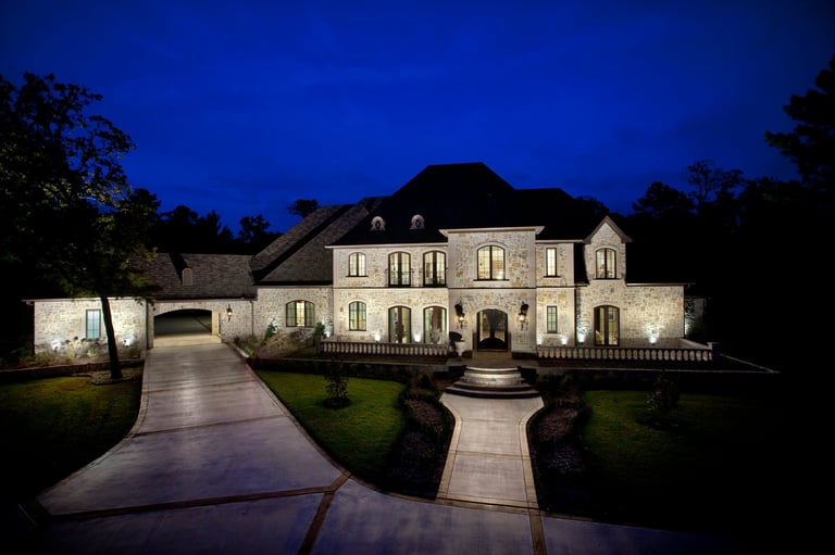 Luxurious mansion illuminated at dusk with manicured lawn, curved driveway, and mature trees framing the elegant stone and brick estate