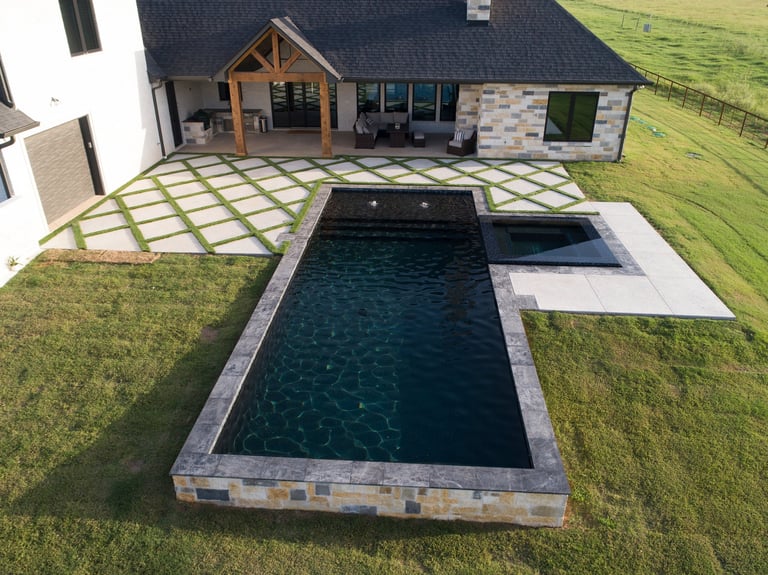 Aerial view of modern home with rectangular dark infinity pool, concrete patio, and manicured lawn
