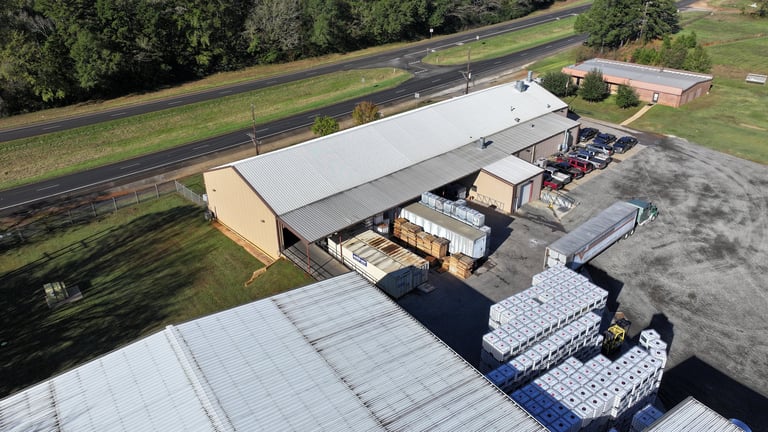 Aerial view of a large warehouse facility with white roof, loading dock with stacked pallets, and a rural road nearby