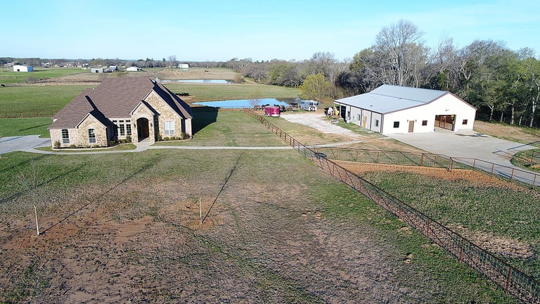 Aerial view of a rural farm property with two houses, fenced pastures, a pond, and trees in the background