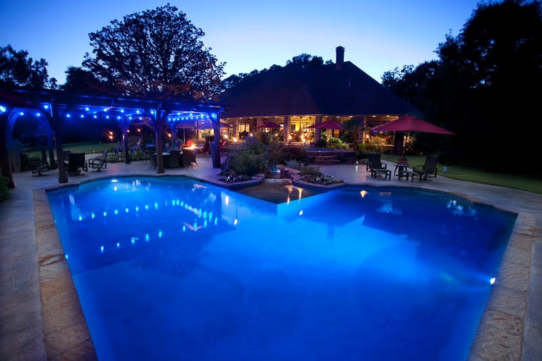 Illuminated blue swimming pool at night with glowing patio, lit pergola, and stone house with warm lighting in background