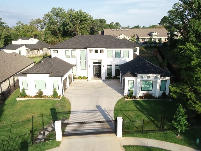 Modern white multi-level mansion with circular driveway, black metal gates, and manicured lawn surrounded by trees