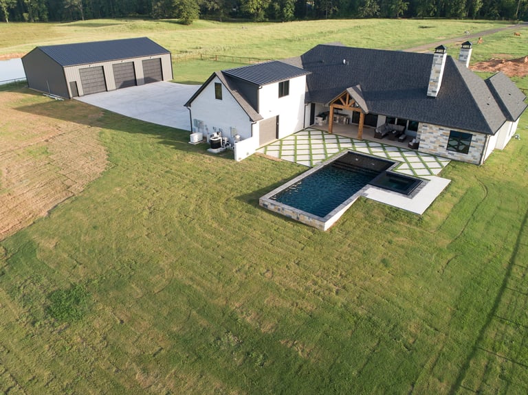 Aerial view of modern farmhouse with attached garage, pool, and hot tub on expansive green property
