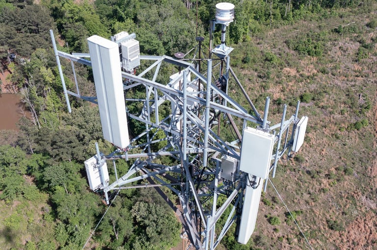 Aerial view of a cell tower with white antennas and equipment mounted on a metal lattice structure surrounded by trees and vegetation.