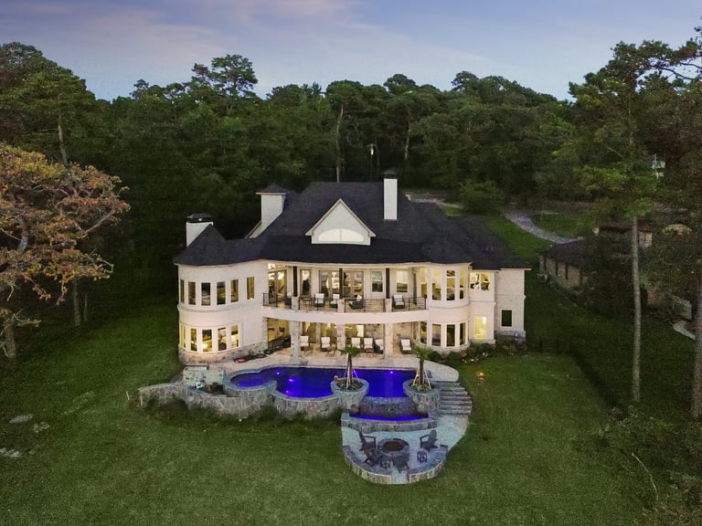 Aerial view of a large cream-colored mansion with black roof, illuminated windows, blue pool, and manicured lawn surrounded by trees at dusk.