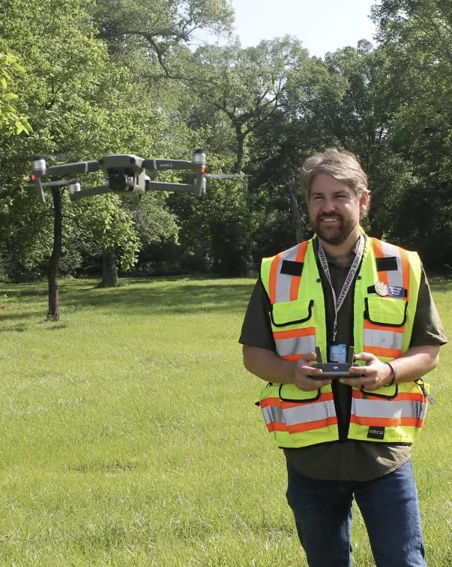 Man in safety vest holding drone controller in grassy field with trees and suspended drone rig in background