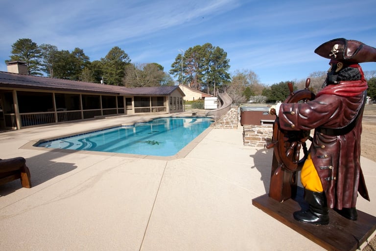 Outdoor swimming pool facility with a covered structure, wooden statue of a pirate figure in foreground, surrounded by trees and clear blue sky