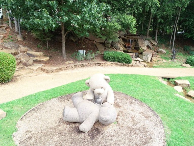Stone sculpture of a sitting bear on a sandy area surrounded by green grass and trees in a garden setting