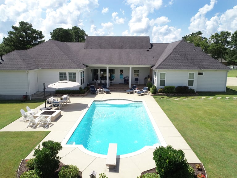 Aerial view of a modern house with a rectangular swimming pool, patio furniture, and manicured lawn surrounded by trees