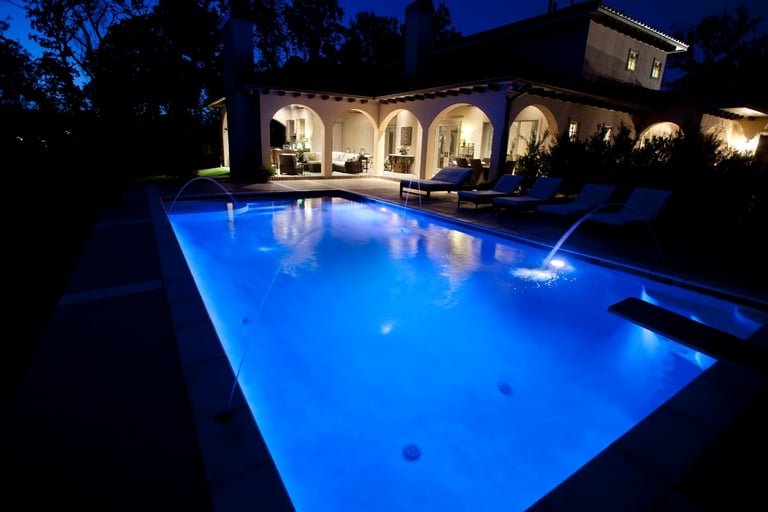 Illuminated rectangular swimming pool with blue LED lighting at night, adjacent to a Mediterranean-style villa with lit arches and trees in background