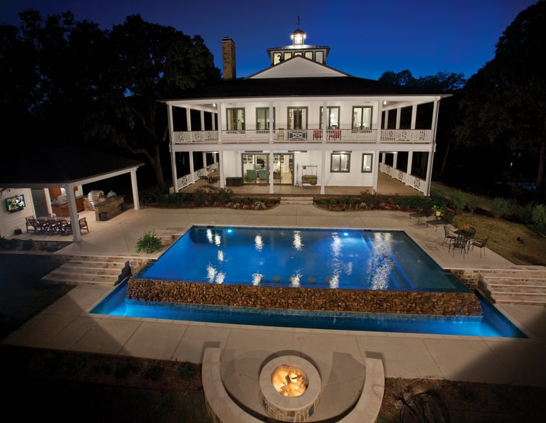 Elegant white mansion with octagonal cupola overlooking illuminated swimming pool at dusk, surrounded by manicured landscaping and trees
