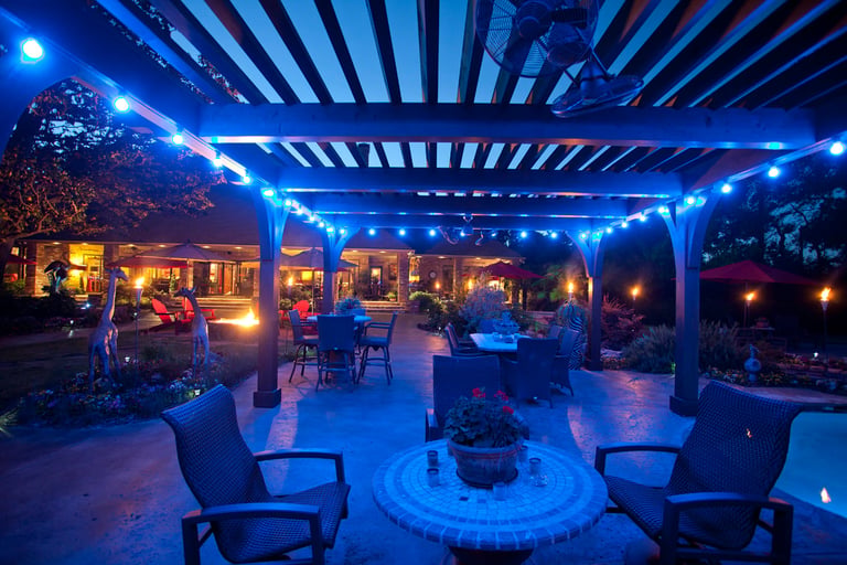 Modern patio with blue LED pergola lighting, fire pit, lounge chairs, and house visible in background at dusk