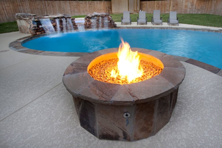 Outdoor fire pit with active flame surrounded by pool decking, with a blue swimming pool and lounge chairs visible in the background