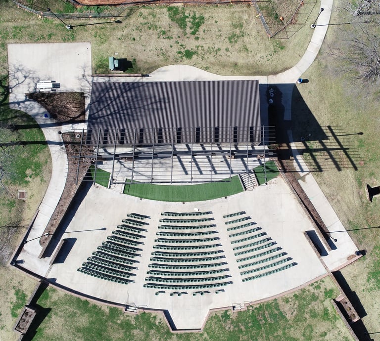 Aerial view of an outdoor amphitheater with a stage building and curved seating arrangement surrounded by green lawn