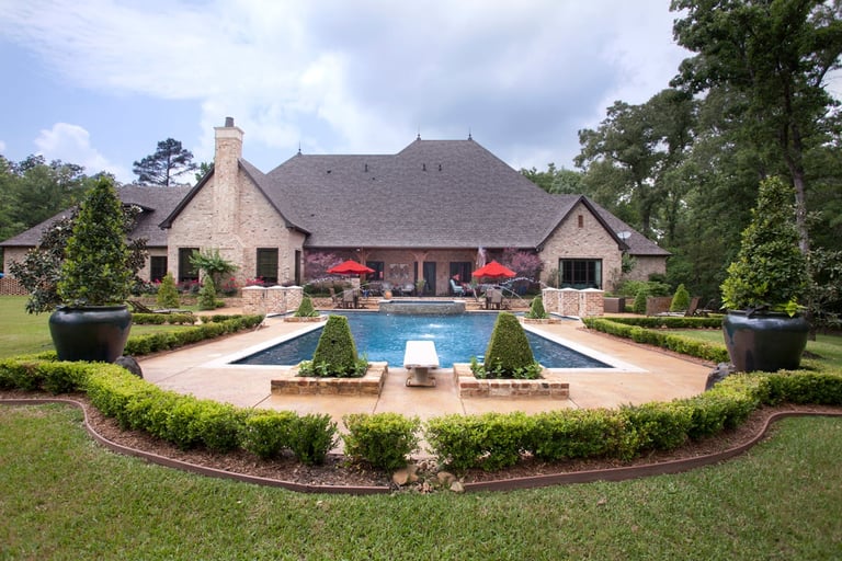 Luxury home with slate roof and stone exterior featuring a rectangular pool, lounge chairs, manicured gardens, and surrounding landscaping