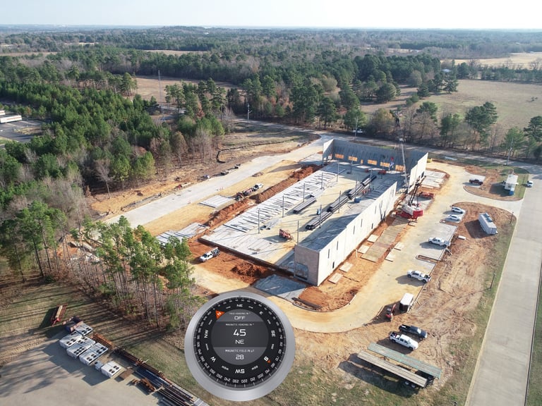 Aerial view of industrial construction site with large warehouse building, compass overlay, and surrounding forest and fields
