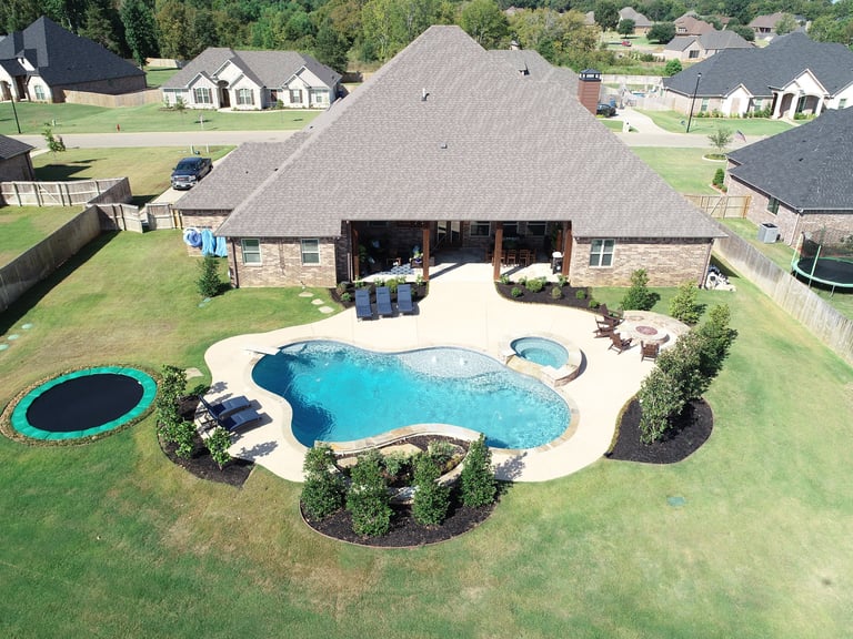 Aerial view of a luxury home with a large backyard pool, hot tub, and trampoline surrounded by manicured lawn