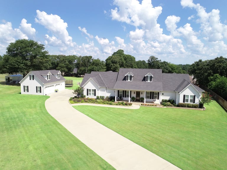Aerial view of a two-story suburban home with a curved driveway, manicured lawn, and surrounding trees under blue sky.