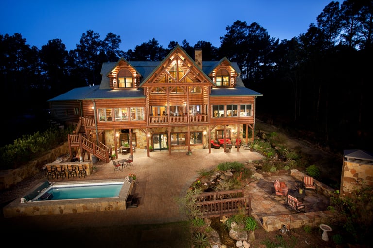 Large two-story wooden house illuminated at dusk with pool and landscaping in wooded setting
