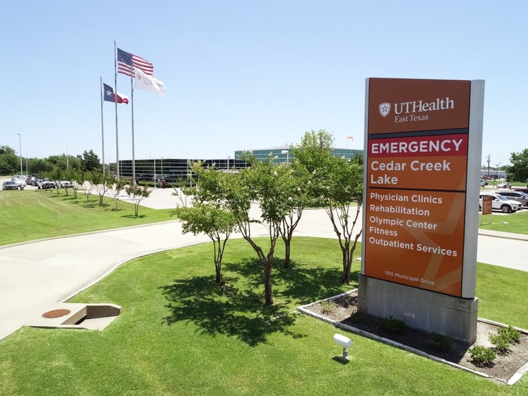 UTHealth Emergency Cedar Creek Lake facility entrance with brown directional sign, flagpoles with American flag, and green lawn