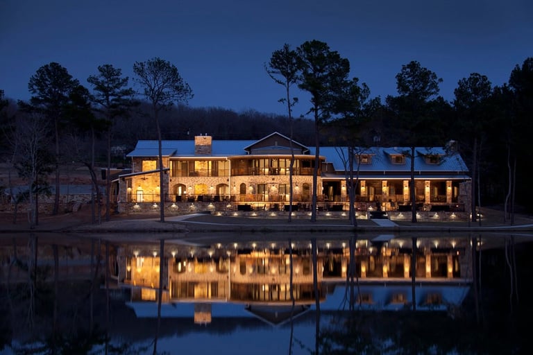 Illuminated two-story house reflected in calm water at twilight, surrounded by bare trees