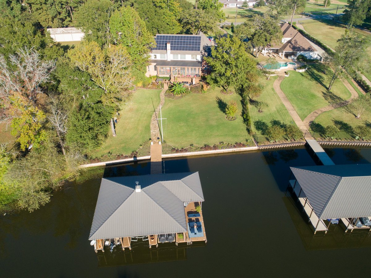 Aerial view of waterfront property with boathouse, residential home with solar panels, and manicured grounds along a calm river