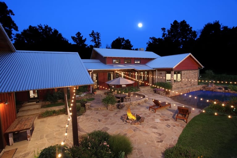 Modern rustic farm complex at twilight with metal-roofed buildings, illuminated courtyard, fire pit, and landscaping under a moonlit sky