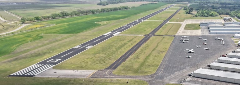 Aerial view of airport runway with taxiways, surrounding green fields, and terminal buildings on the right side