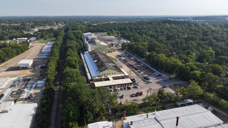 Aerial view of industrial facility with blue-roofed buildings, warehouses, and parking areas surrounded by dense green trees