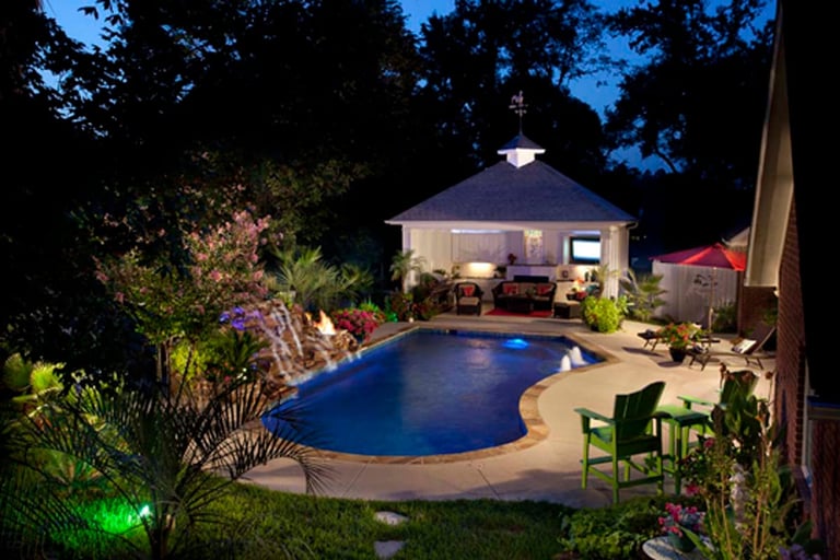Nighttime backyard with illuminated pool, poolhouse, lush landscaping, and patio seating area