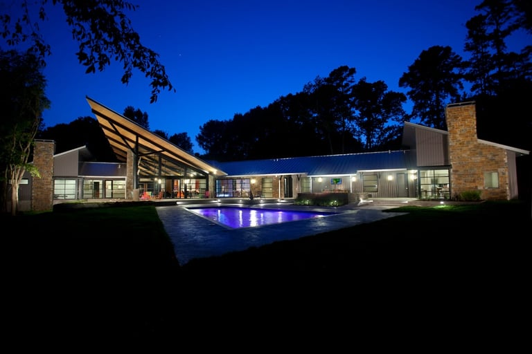 Modern house at night with illuminated pool, featuring contemporary architecture, floor-to-ceiling windows, and landscaping against a twilight sky.