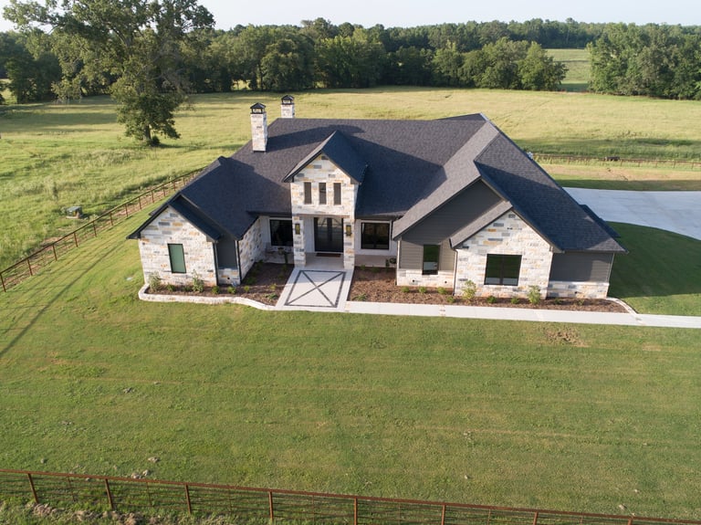 Aerial view of a modern ranch-style home with dark roof situated on expansive green grounds with a pond and surrounding farmland