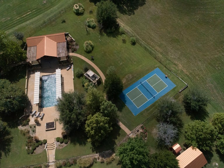 Aerial view of a residential property with a terracotta-roofed house, swimming pool, and blue tennis court surrounded by green lawn and trees.