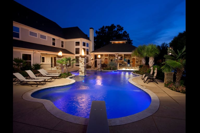 Luxury home with illuminated blue resort-style pool, lounge chairs, and tropical landscaping at twilight