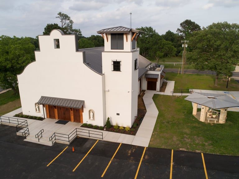 Aerial view of a modern white church building with metal roof, parking lot, and green lawn surrounded by trees