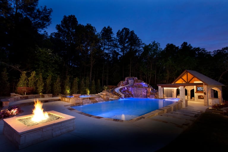 Nighttime backyard with illuminated swimming pool, waterslide, fire pit, and pool house surrounded by tall trees