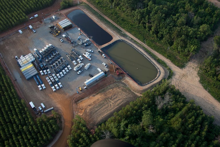 Aerial view of an industrial facility with storage tanks, equipment, and a large retention pond surrounded by forest and farmland