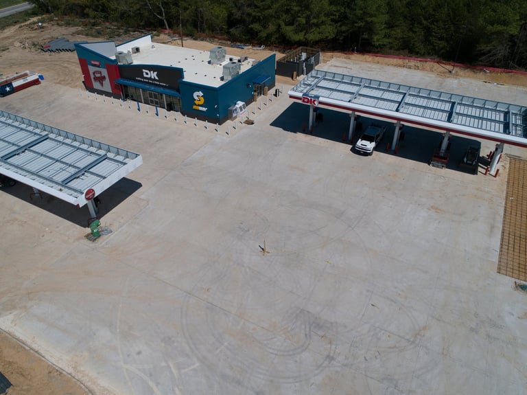 Aerial view of an industrial compound with concrete courtyard, blue warehouse building, solar panels, and black structures surrounded by forest.