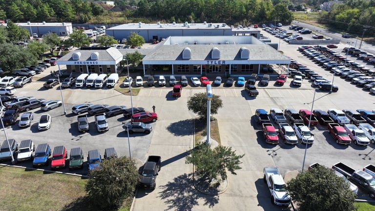 Aerial view of a large car dealership lot with numerous vehicles parked in rows and a showroom building with blue roof