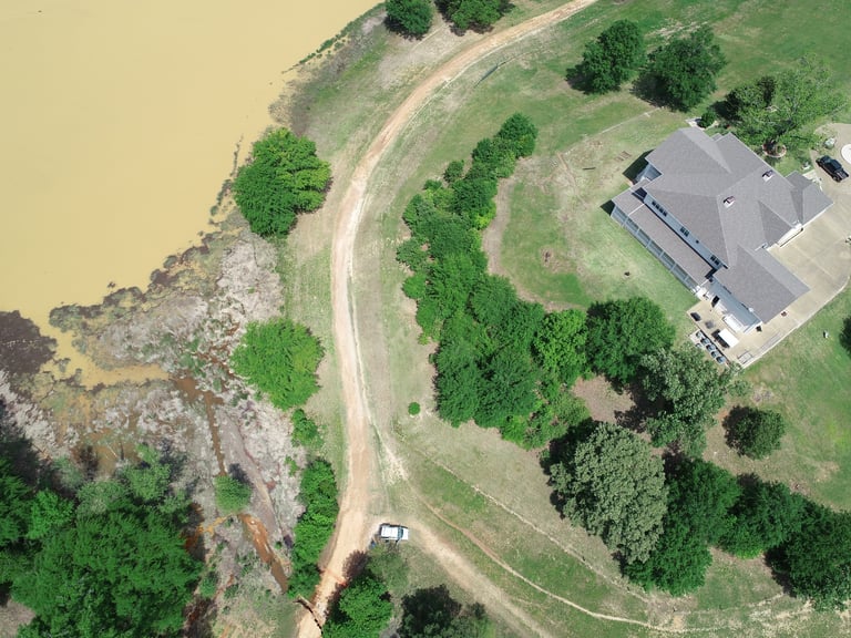 Aerial view of a property with a gray-roofed building, winding dirt driveway, green trees, and a yellowish pond or water body on the left