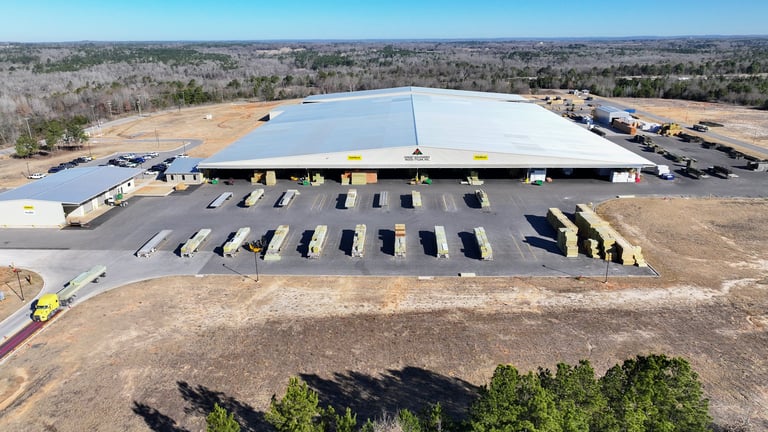 Aerial view of a large industrial warehouse with blue metal roof and parking lot with yellow line markings in a sparse desert landscape.